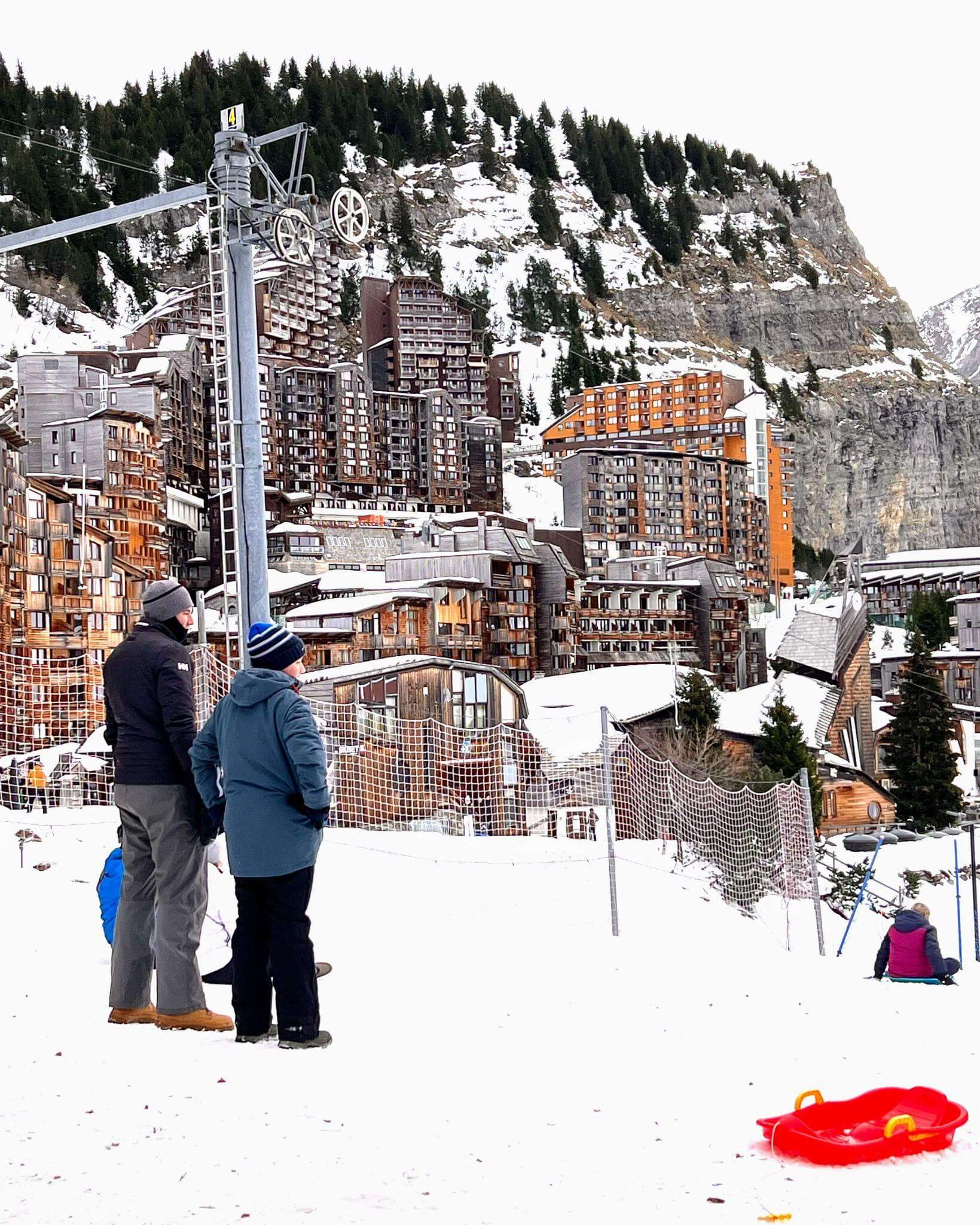 Family watching so sledding down a slope in the middle of Avoriaz ski resort, snow capped mountains and unusual shapes of the resort buildings in the background