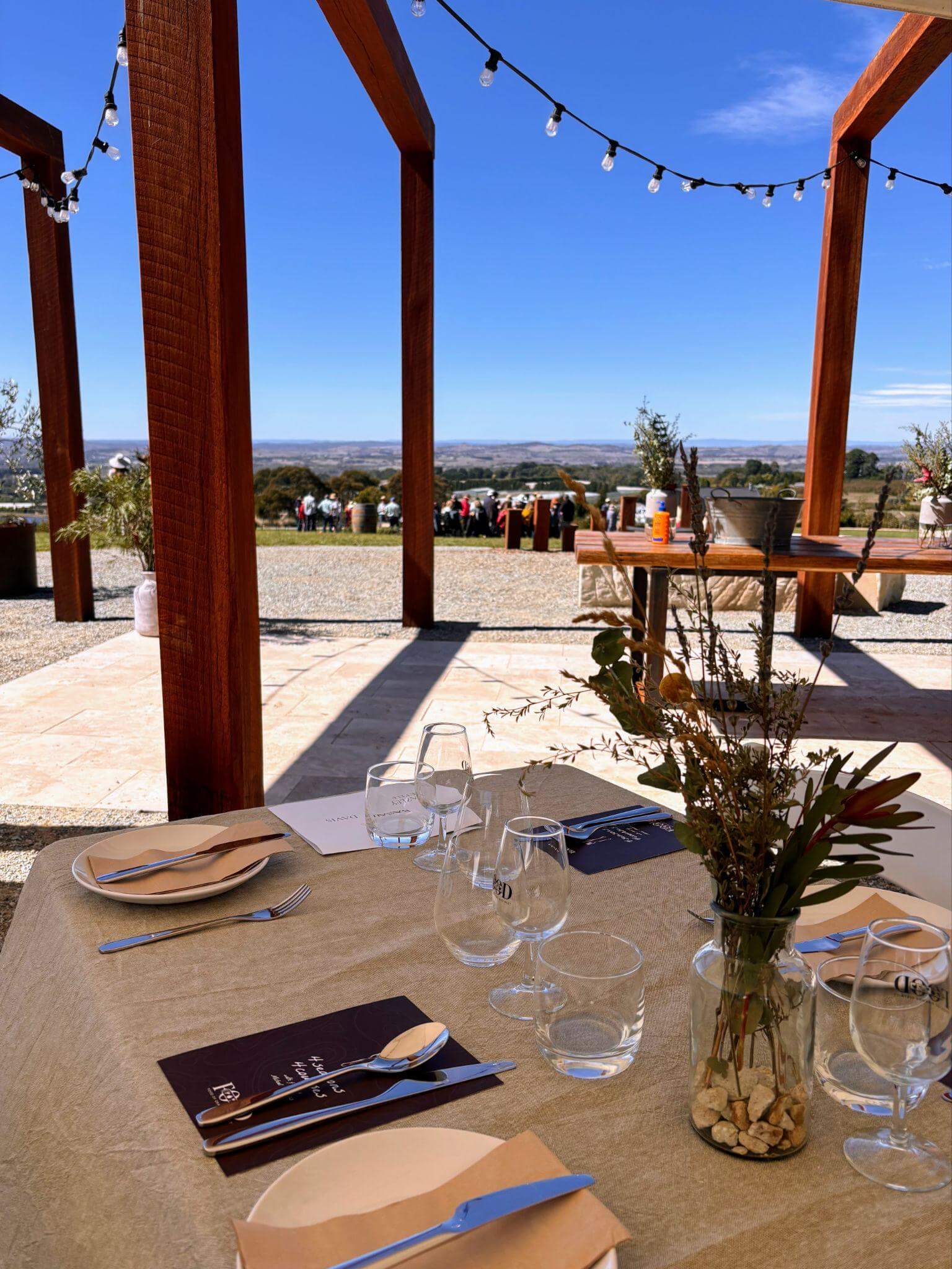 Long table with linen tablecloth and set with native flowers overlooking a group of party goers and panoramic views over the town of orange in the background