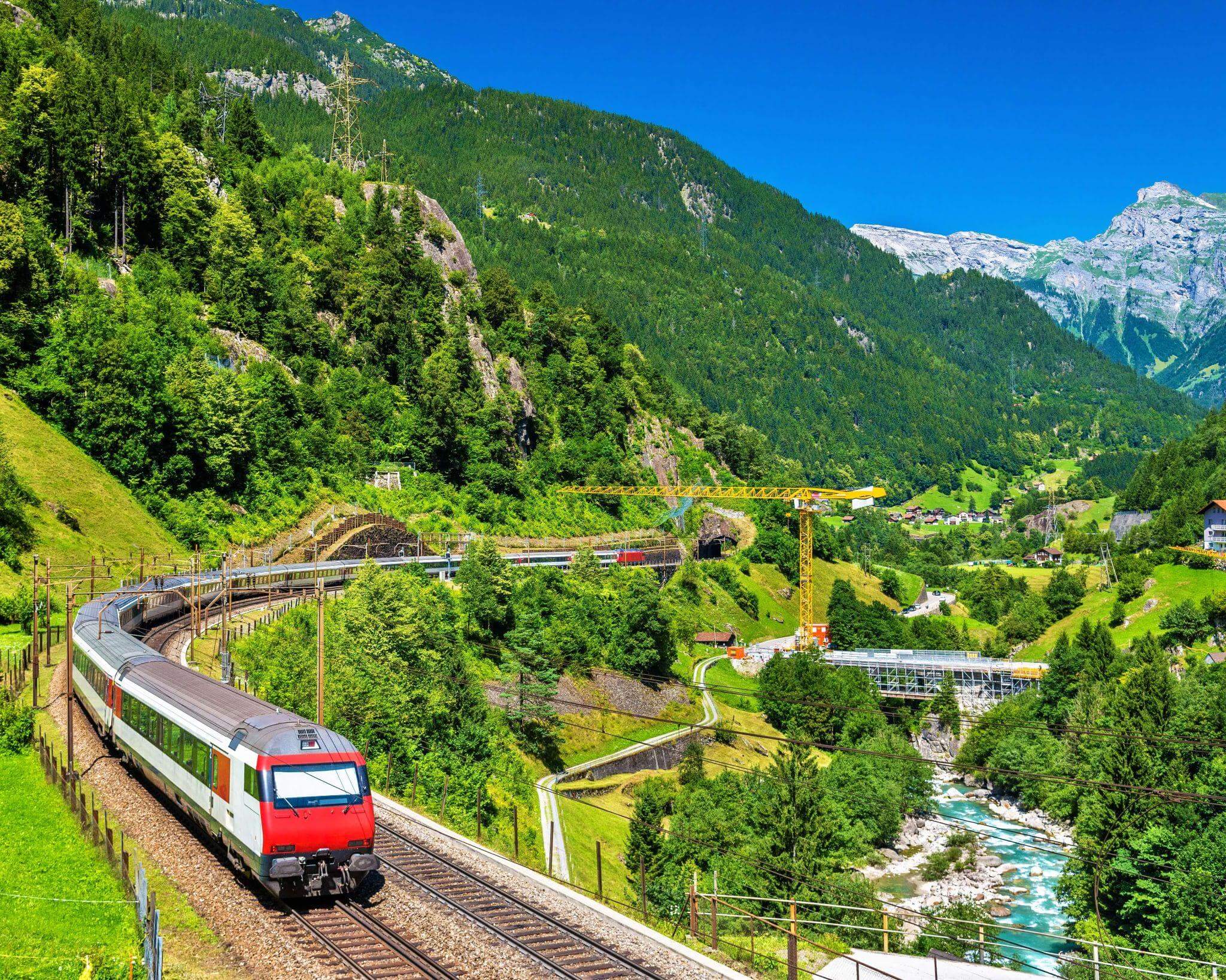 red and white train travelling through the swiss alps in summer with bright green trees on the hills and turquoise blue river in the valley below