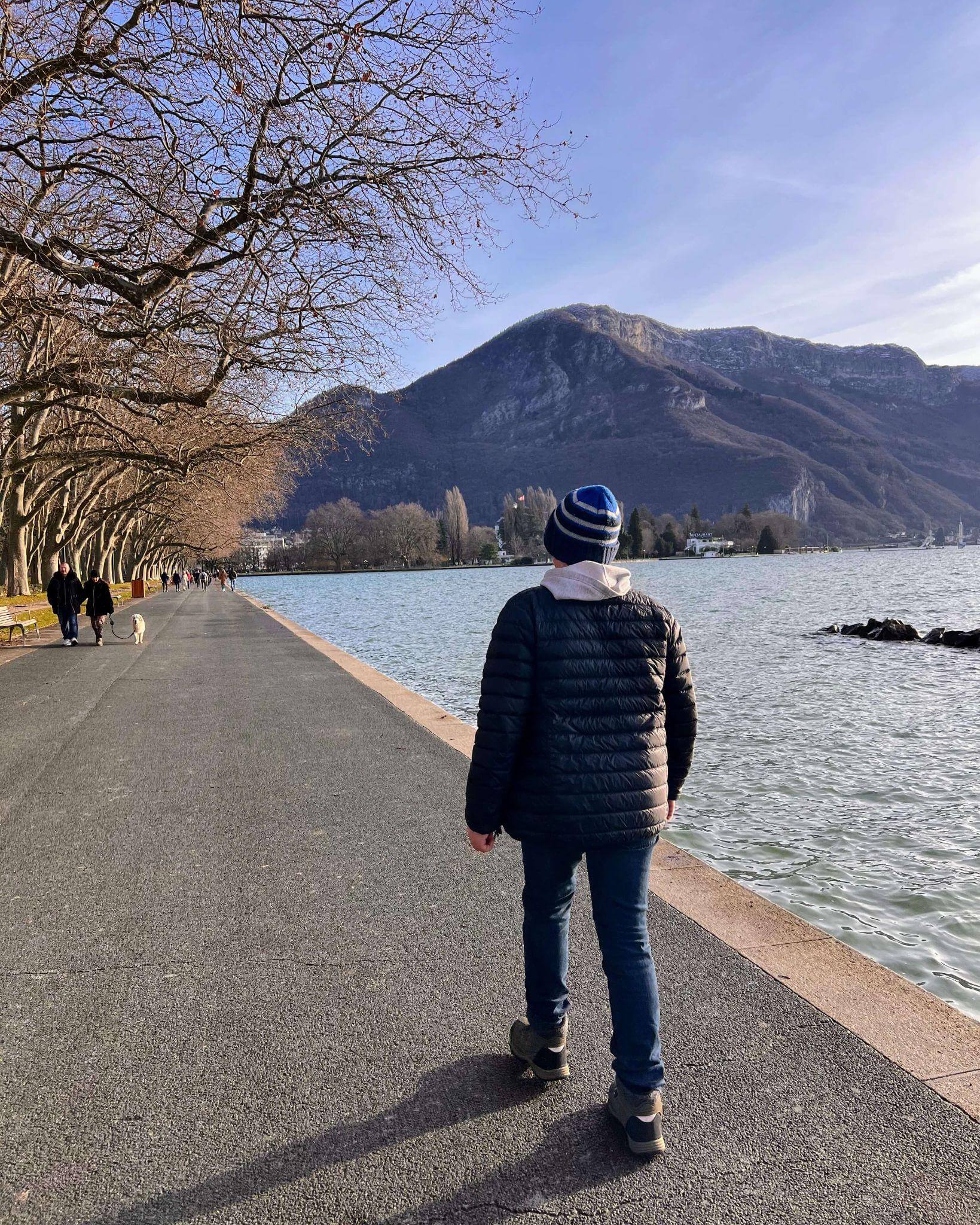 Teenager walking alongside Lake Annecy in France, it's winter with the teen wearing a puffer jacket and beanie and the trees are bare