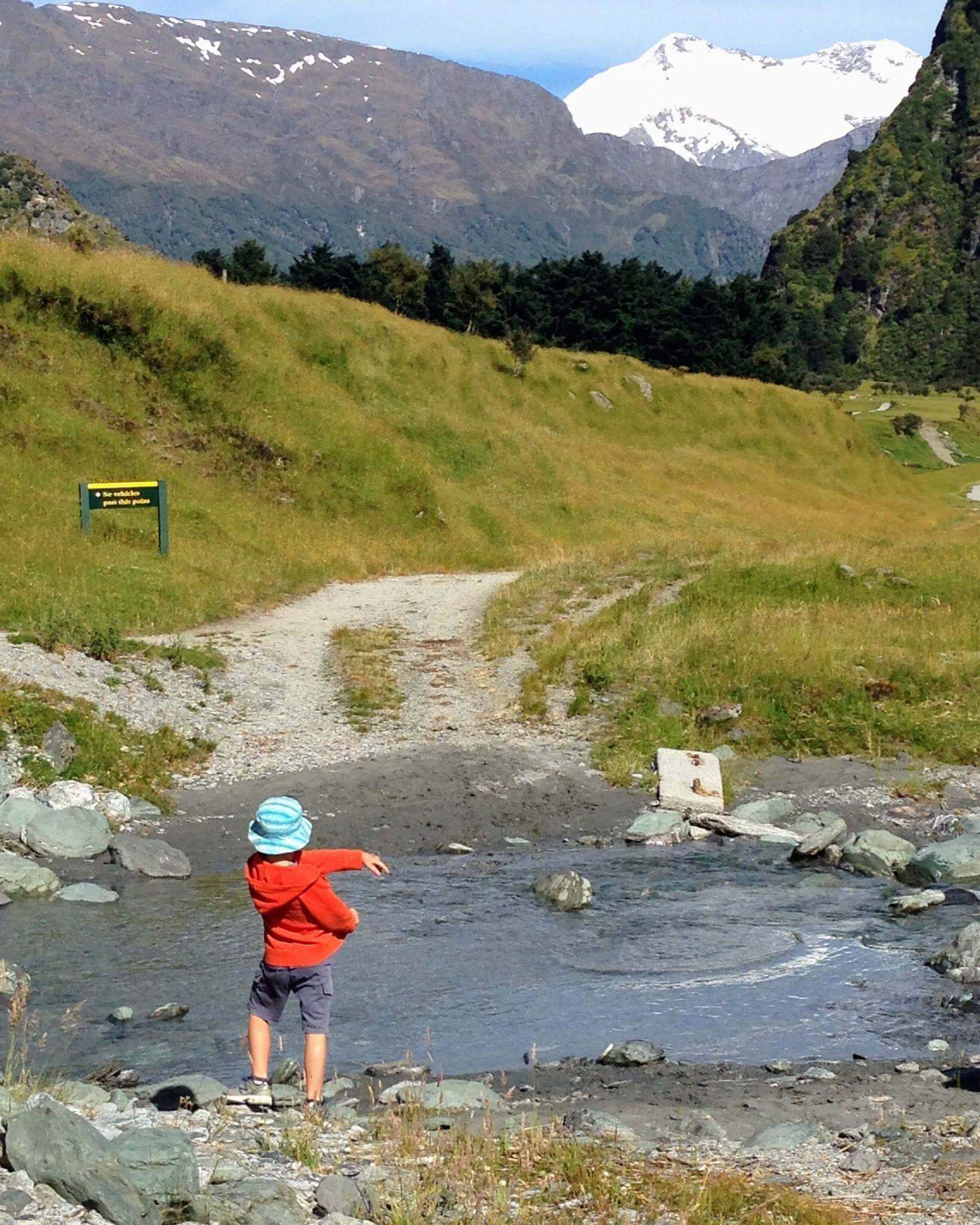 Young boy skimming a rock into a clear mountain stream with snow capped mountains in the background in New Zealand