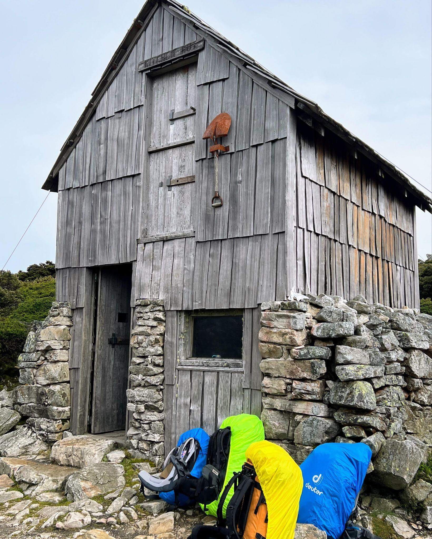 old shingle rest hut with 4 colourful backpacks on the outside
