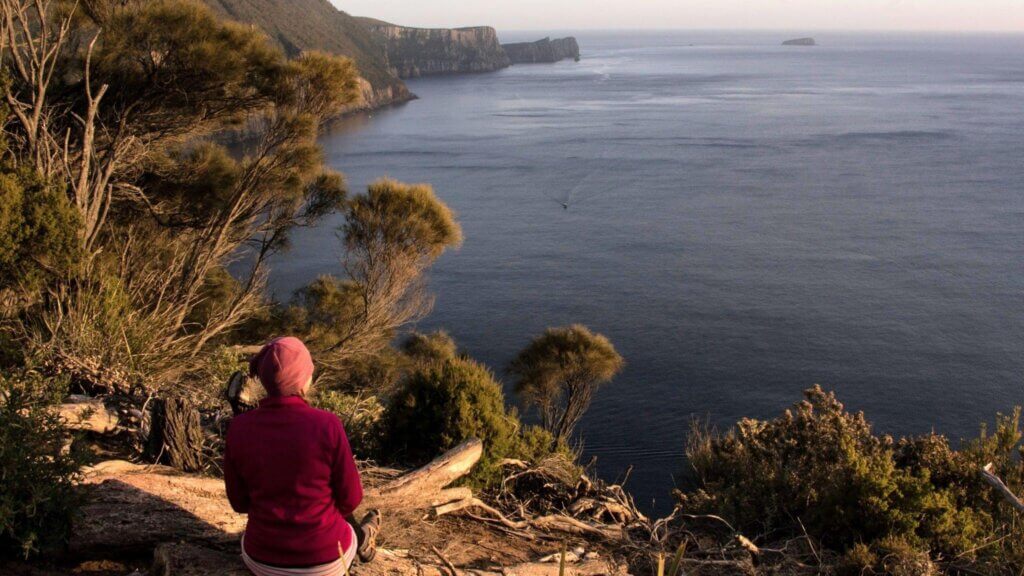 rachel-reinke-looking-out-over-an-expansive-ocean Rachel Reinke looking out over an expansive ocean at sunset from a high lookout point with cape pillar in the distance