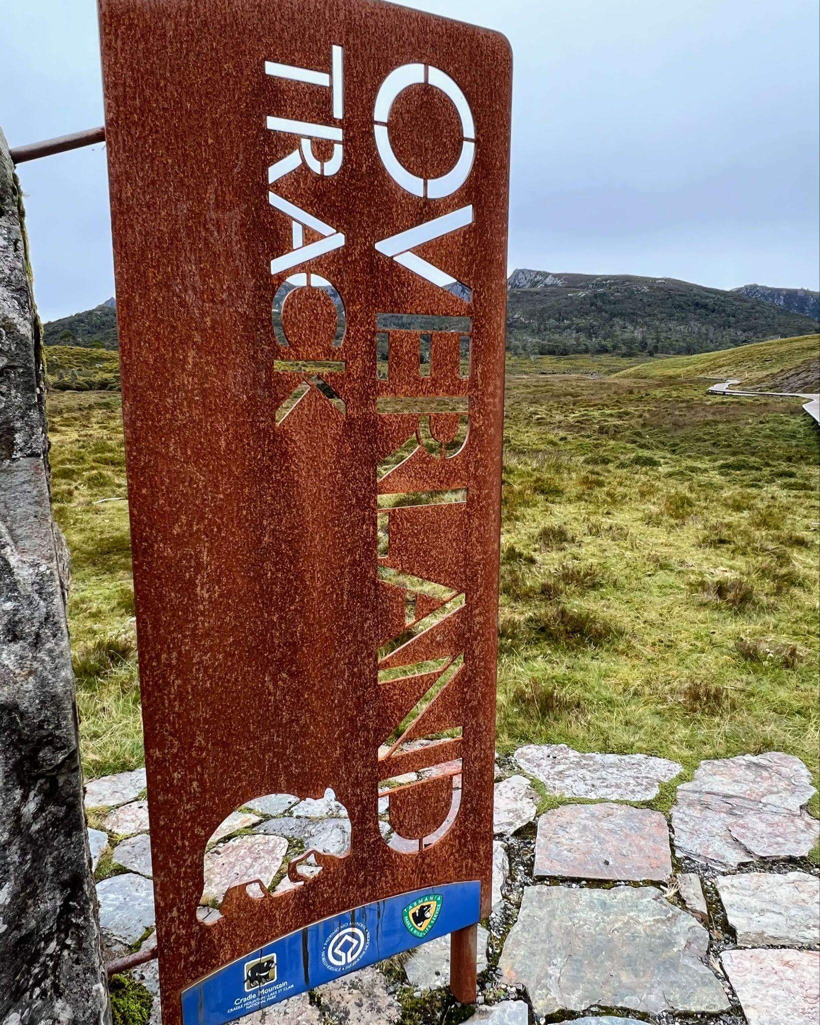 Signpost showing the official starter of the overland track with boardwalk and panoramic view of the mountains in the background