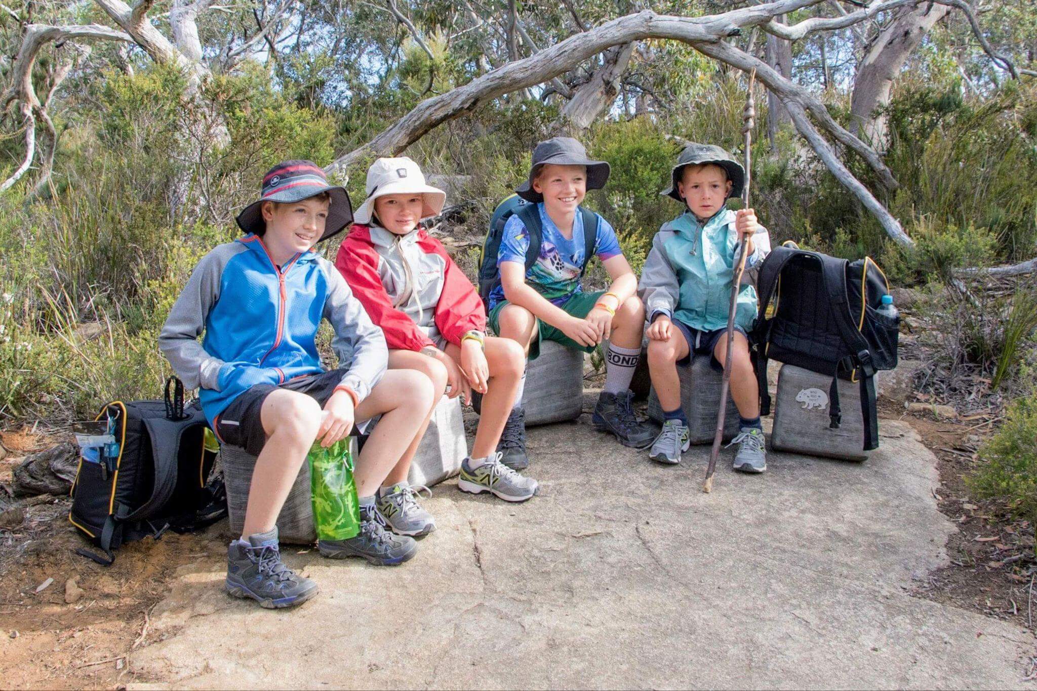 Four children taking a rest break during multi-day hiking with kids, sitting on log with backpacks and wearing sun hats in Australian bush setting