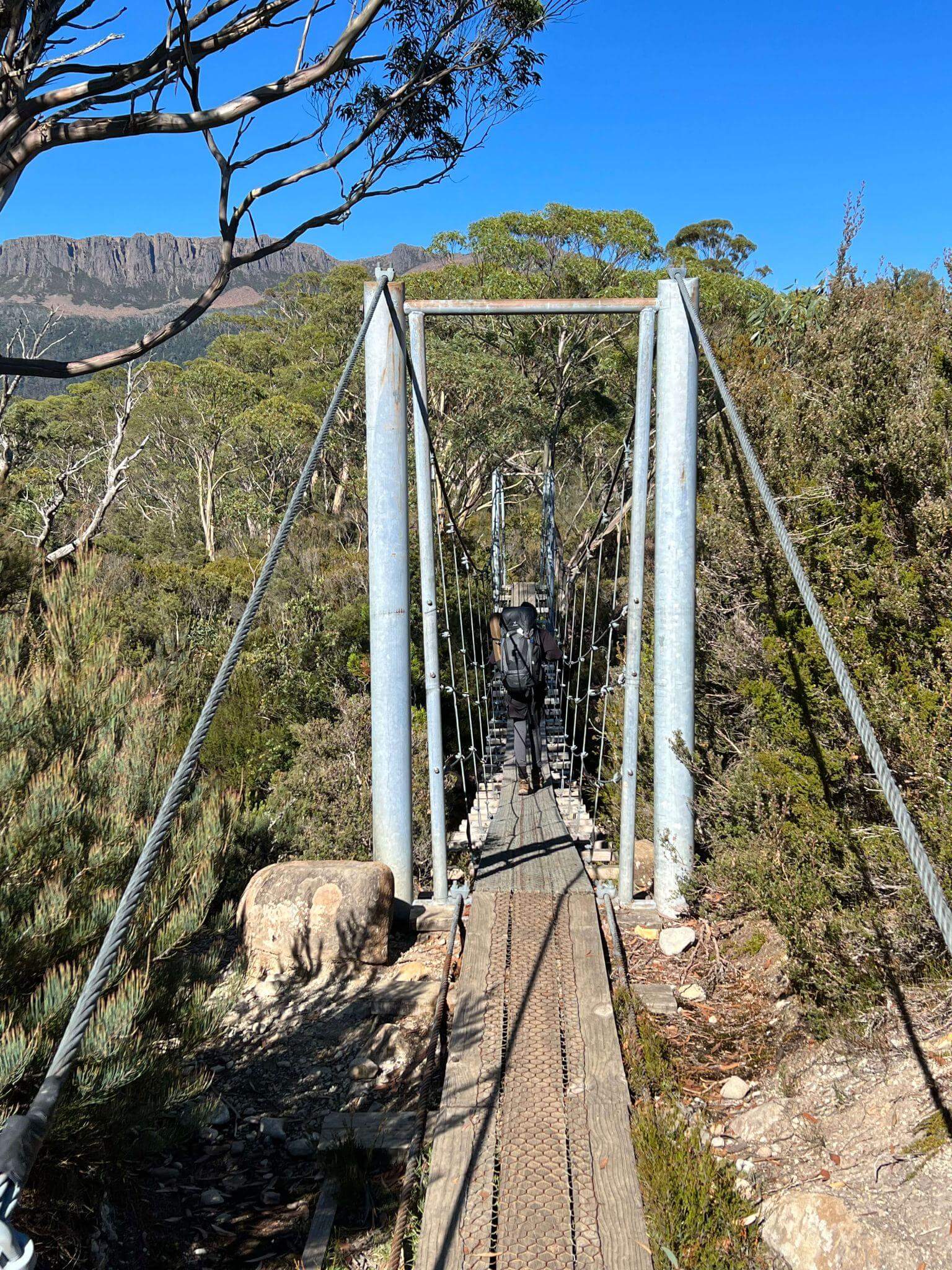 Hiker crossing wooden suspension bridge over gorge during multi-day walk with full backpack in Australian wilderness