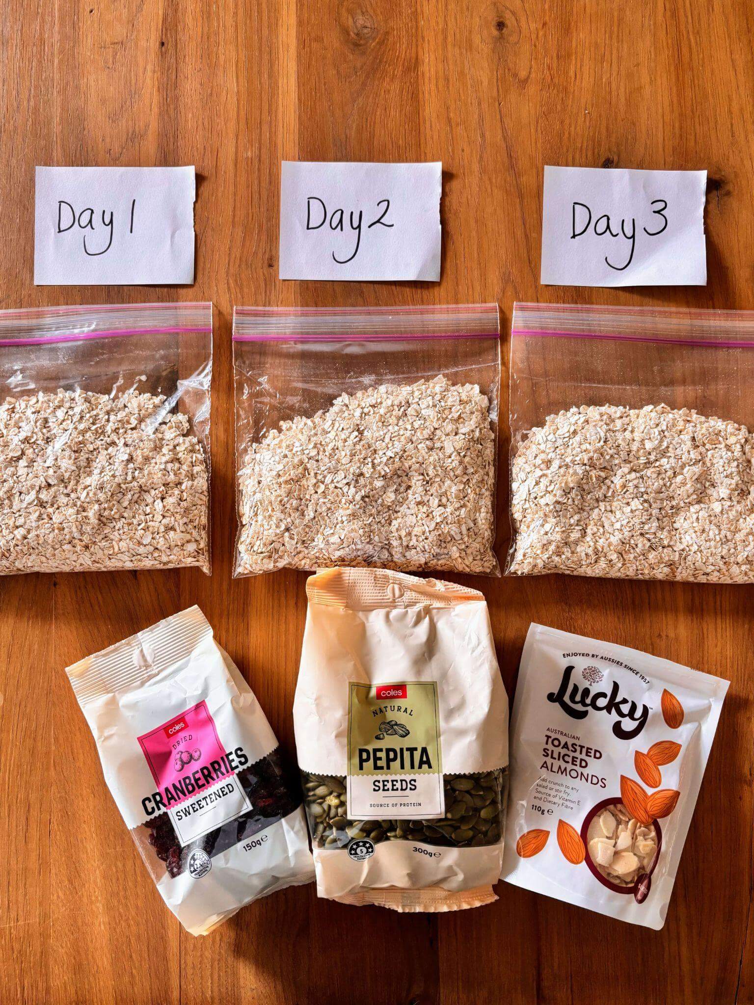 Multi-day hiking food organization showing three ziplock bags labelled Day 1, Day 2, and Day 3 filled with oats, alongside cranberries, pepita seeds, and sliced almonds for trail breakfast preparation