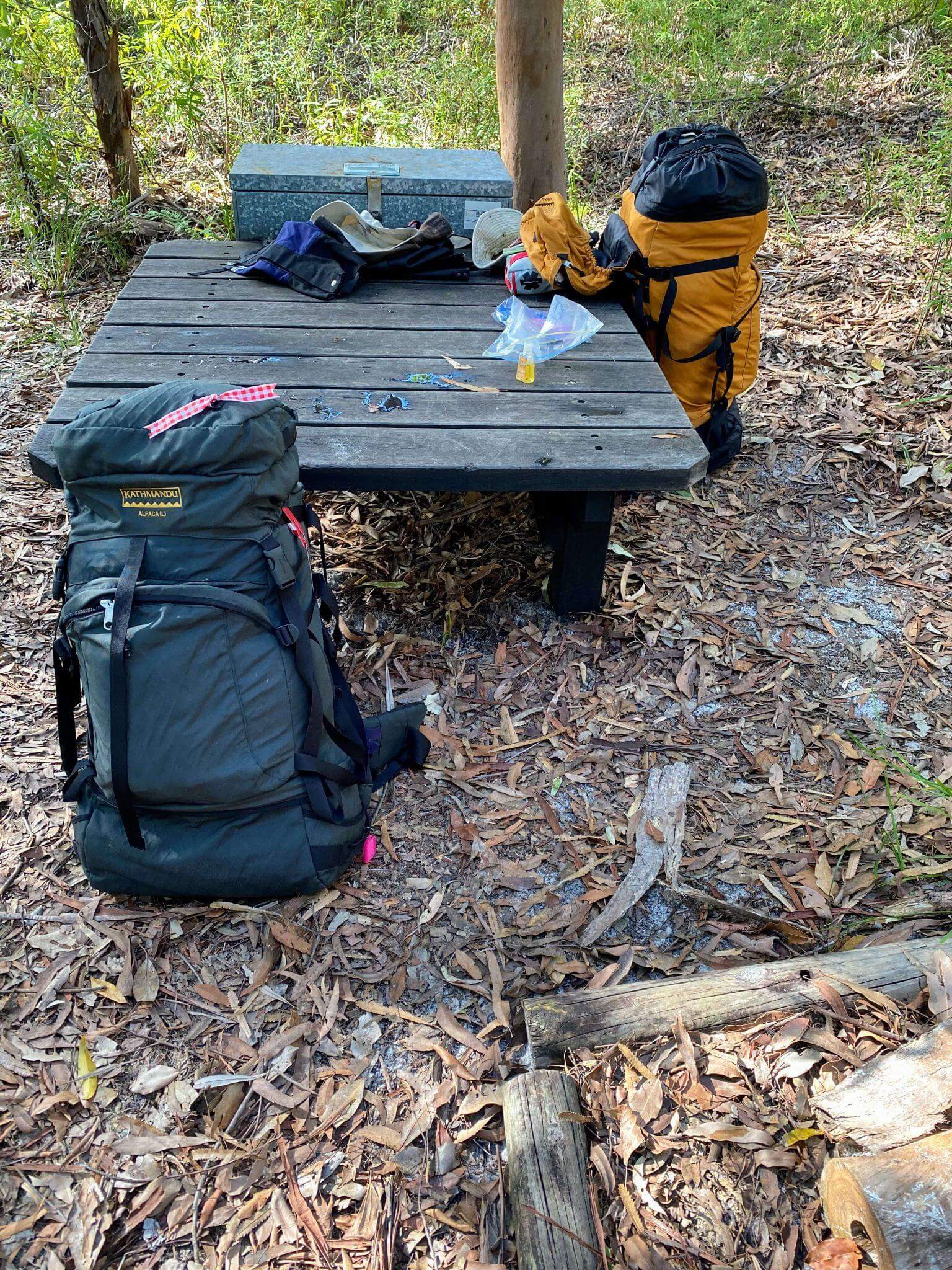 Multi-day hiking gear and backpack spread on wooden table at campsite showing essential equipment and lunch break setup
