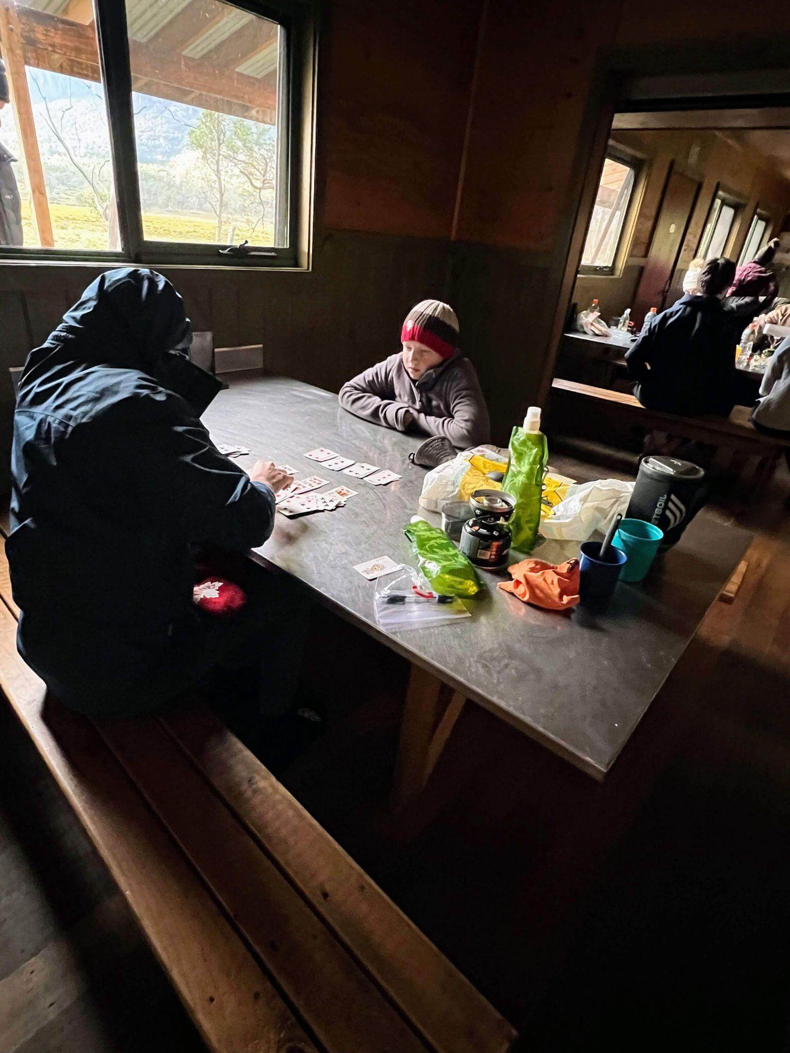 Multi-day hiking meal preparation inside mountain hut showing hikers using camping stove and organizing hiking food supplies on wooden table