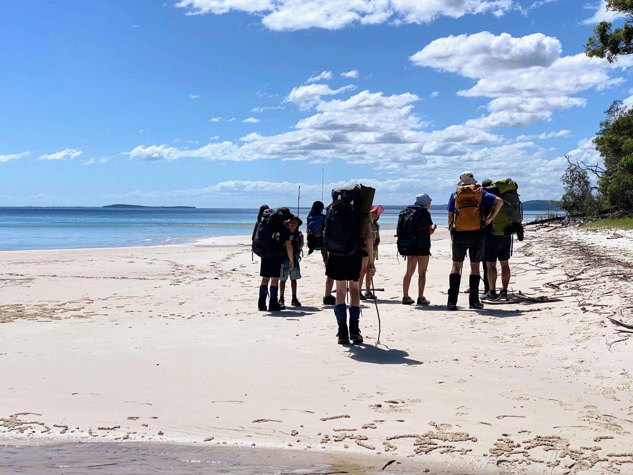 Multi-generational hiking group with children carrying backpacks walking along pristine beach during multi-day coastal hiking adventure under blue sky