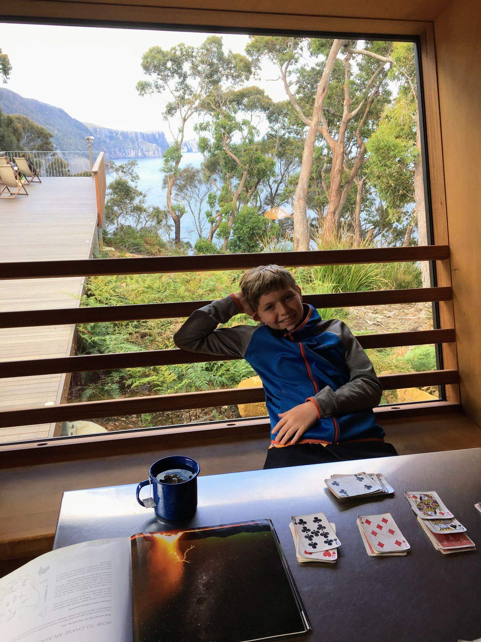 Young boy playing cards at table inside hiking hut during multi-day hiking with kids adventure, with scenic lake and forest views through window